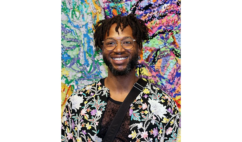 Black man with glasses, short hair, and a beard, wearing a colorful floral print jacket, smiling into the camera photographed against a colorful and abstract backdrop